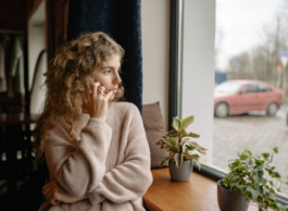 A person holds a phone to their ear while leaning on a counter with houseplants and looking out the window at a parking lot.