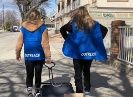 Two people in blue vests that say Outreach walk down a city sidewalk on a sunny day.