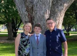 Two smiling adults in business casual attire stand outside in front of a tree on either side of a youth wearing a suit and tie.