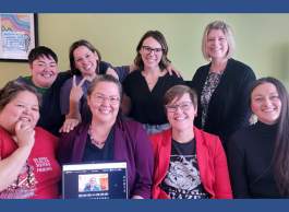8 people in two rows of four, one of which holds a laptop of person connected virtually, cheerfully pose for a picture.