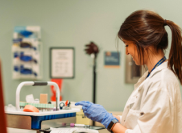 A person wearing scrubs and medical gloves smiles while labelling lab samples next to a collection chair.