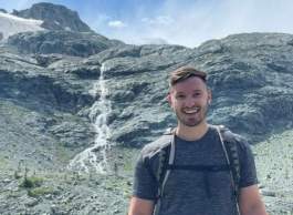Person hiking in grey t-shirt standing in front of a rockface with a waterfall.