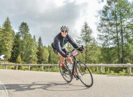 A cyclist rides a road bike around a bend on a paved mountain road lined with trees, with another cyclist visible in the background.