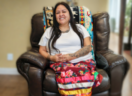 A person with long black hair wears a white shirt and a flower skirt with cultural ribbons sits on a black leather chair indoors and smiles to camera.